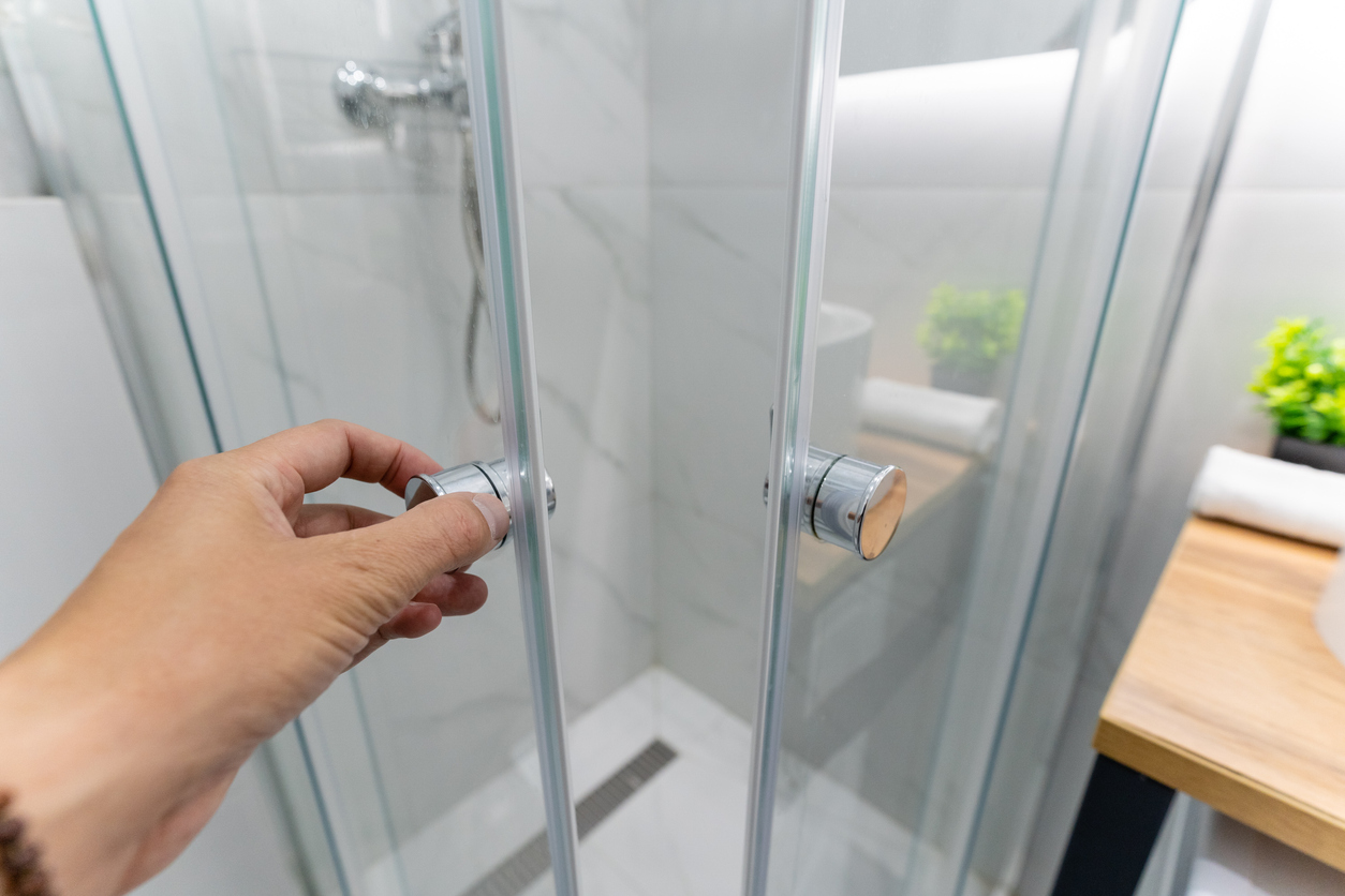 A person in a bathroom is carefully opening a sleek glass shower door, creating an inviting entrance into the refreshing space behind it